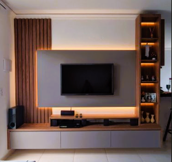 A modern living room entertainment center with a flat-screen TV mounted on a gray panel with warm LED backlighting. To the left is a vertical wood slat accent wall, and to the right, a tall wooden shelving unit with illuminated shelves. Below the TV is a light wood console with gray cabinet doors. A black speaker stands on the left.